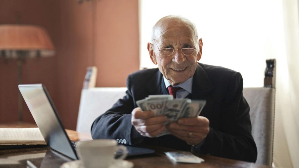 Happy senior businessman counting money while sitting at table with laptop