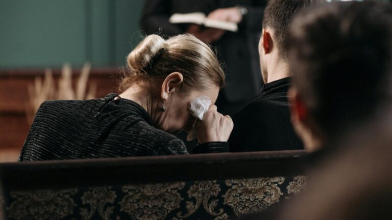 A grieving woman wipes tears during a heartfelt funeral ceremony in a somber indoor setting.