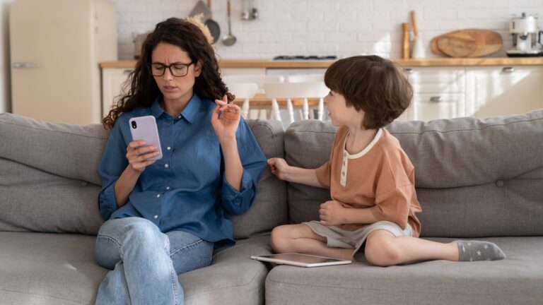 Focused on mobile phone young mother pay no attention on bored small kid son sitting on sofa near