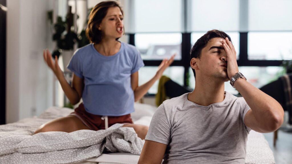 Young man feeling stressed out and having a headache while his girlfriend is arguing with him in the bedroom