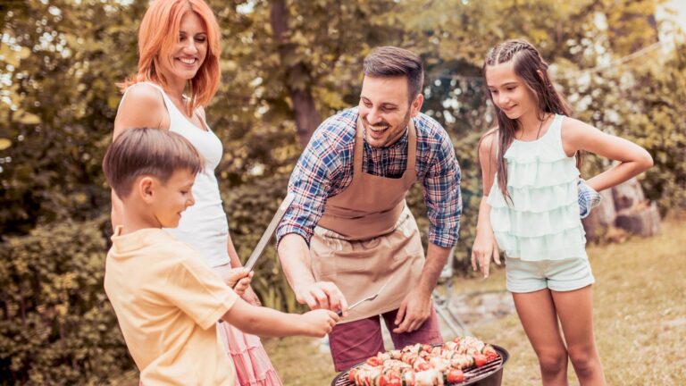 Father teaching son cooking on barbecue with family in background