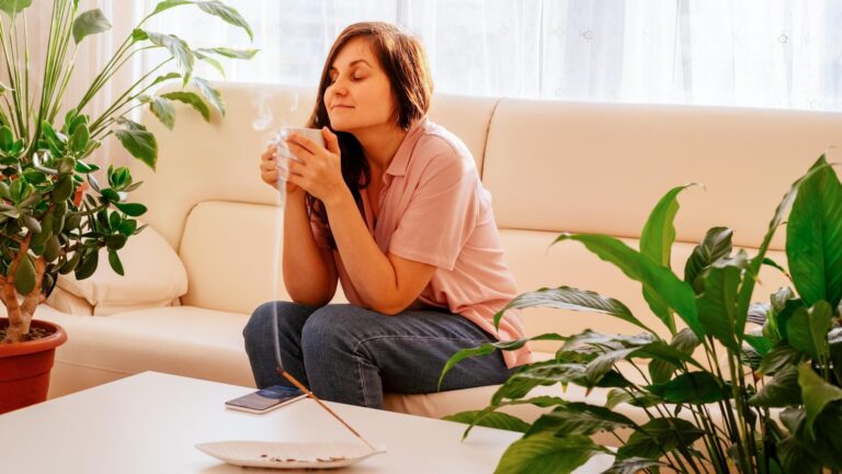 Young beautiful woman is sitting on the sofa drinking coffee and inhaling the fragrance of incense