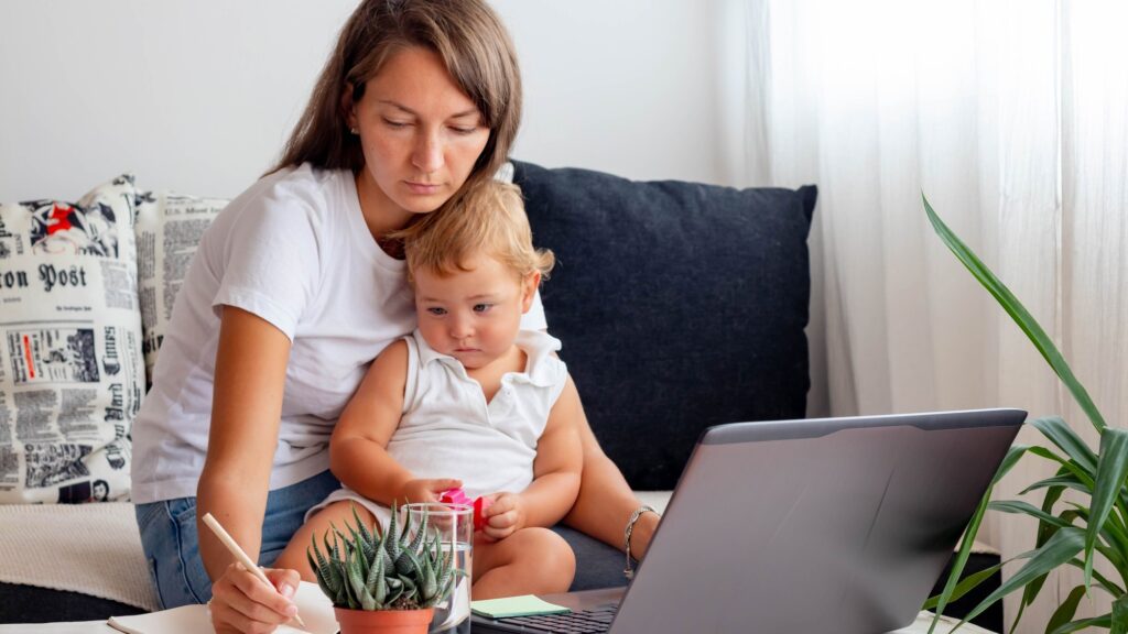 Woman with baby seats close to Laptop with notebook, glass of water on white table in home workspace