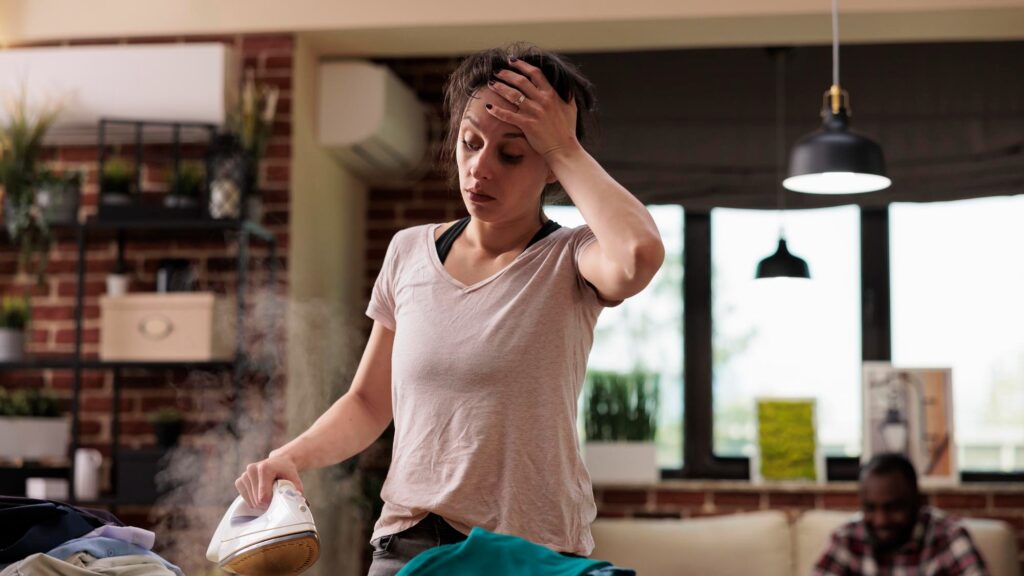 Woman suffering from severe headache ironing clothes while husband relaxes sitting on the sofa using mobile phone. Exhausted wife uncomfortably finishing household chores.