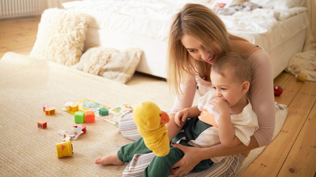 Happy young mother playing and cute little child playing on floor. Blonde female babysitting adorable infant sitting on carpet in bedroom holding yellow duck toy. Motherhood and childcare concept
