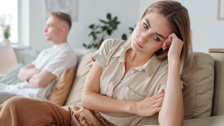 Young upset and lonely wife sitting on couch with her hand by head while expressing offense and misunderstanding during quarrel with husband