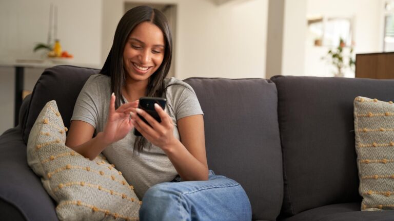 Young biracial woman in casual clothing using mobile phone while sitting on sofa at home