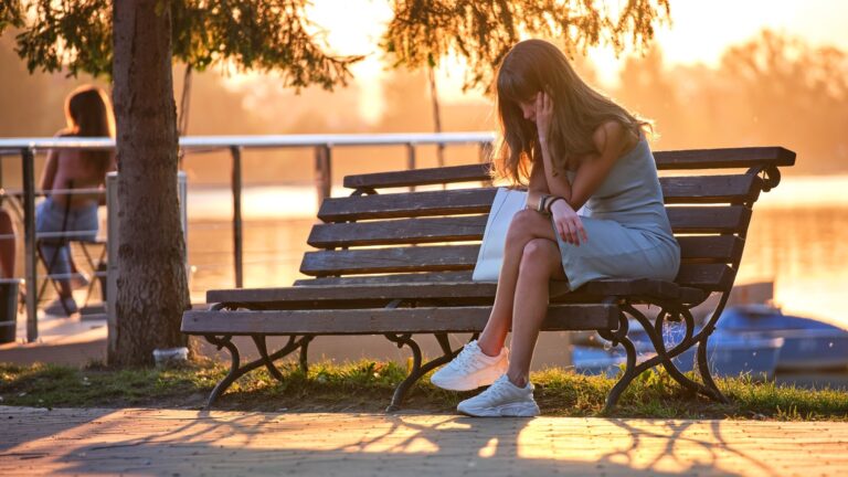 Lonely young woman sitting depressed on lake side bench on warm summer evening. Solitude and relaxation concept.