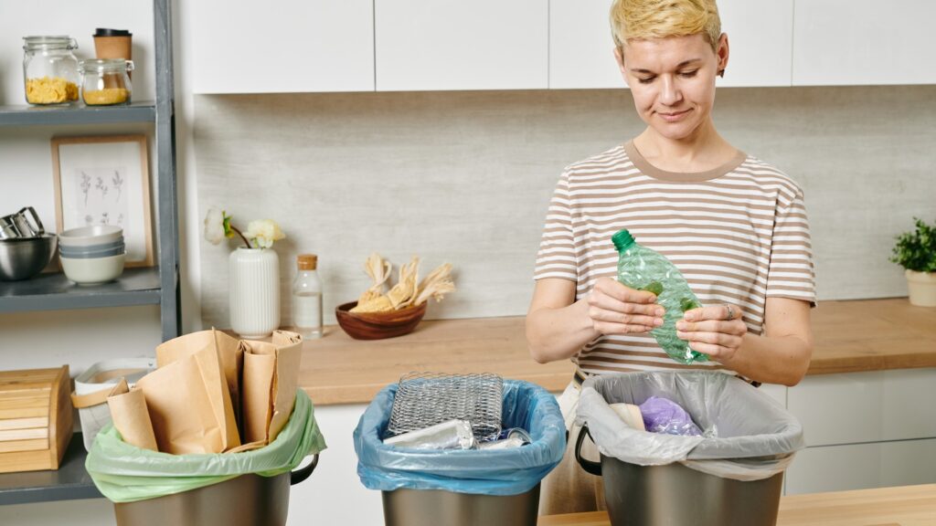 A woman holding plastic bottle over trash bin while sorting waste in the kitchen