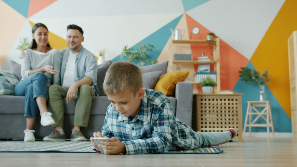 Boy plays on phone while parents watch from couch.