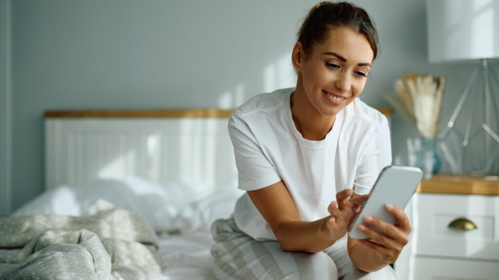 Young happy woman text messaging on cell phone in bedroom