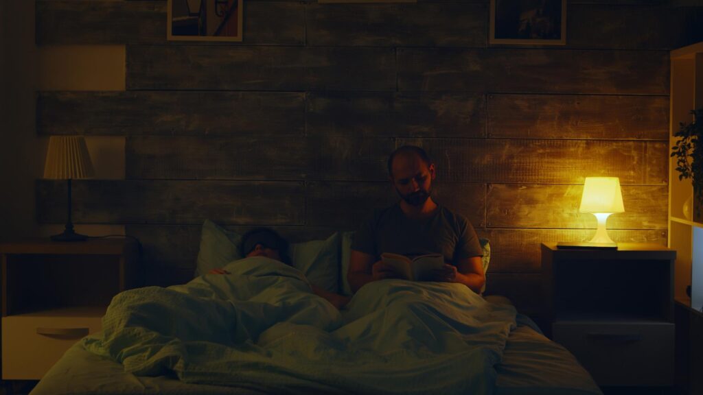 Young man reading a book while his wife is turning the lights off to sleep.