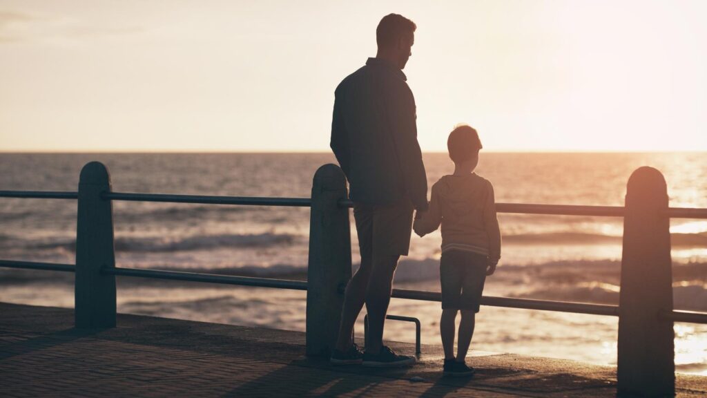 Silhouette sunset and a father holding hands with his son on the promenade at the beach while walking together View family or kids and a man bonding with his young male child outdoor in nature