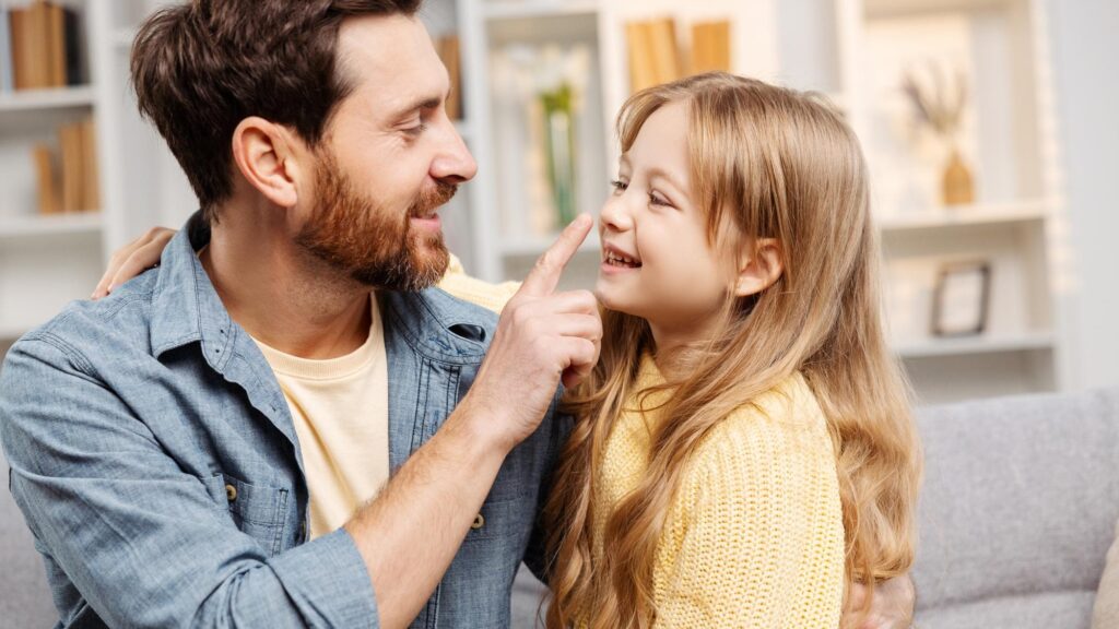 Smiling father lightly touching his young daughters nose Both are sitting on sofa in living room