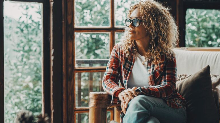Hipster style pretty lady sitting at home on the sofa and looking outside thw windows a green forest woods. Cozy cabiness chalet female people relax lifestyle. Young adult woman enjoy leisure at home