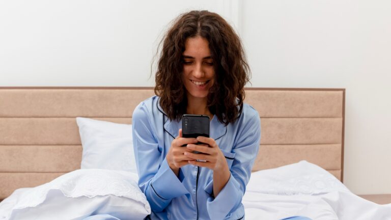 Funny young beautiful woman in blue pajamas sitting on bed using smartphone looking at screen smiling in bedroom interior on light background