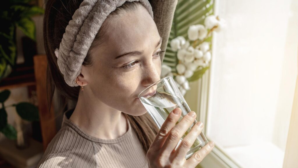 Young woman is standing in front of the window enjoying a sunny morning and drinking clean water from a clear glass Concept of a pleasant start to the day and healthy habits