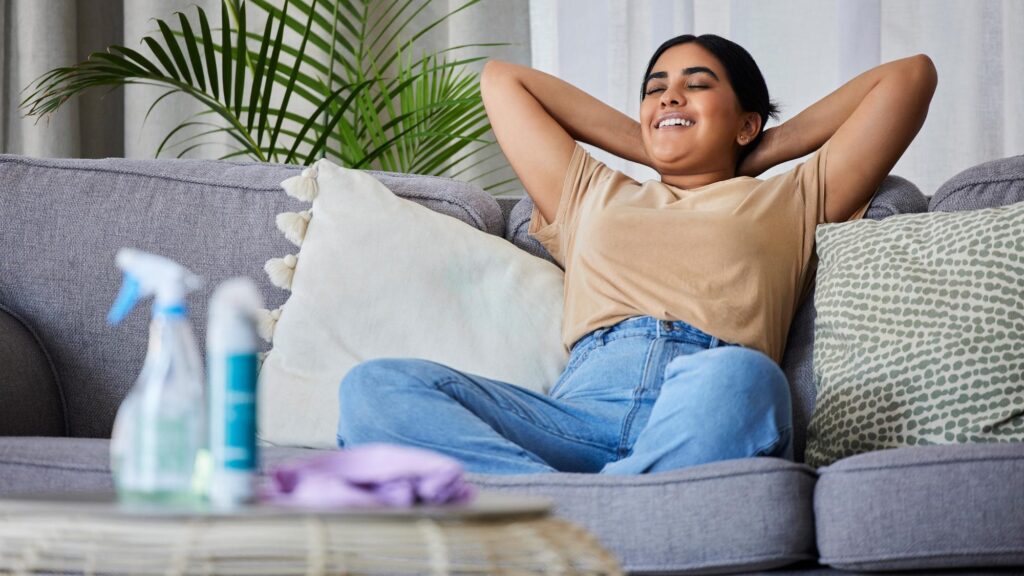 Woman relax and sofa after cleaning house with smile satisfaction and pride in home living room Cleaner rest and happy on lounge couch with spray cloth and hygiene product on table in apartment