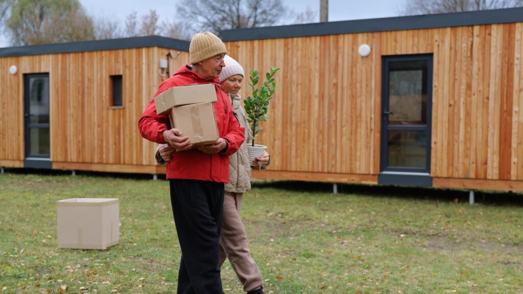 A man and woman walk together holding boxes and a potted plant outside wooden cabins during a cool autumn afternoon showing teamwork and care for their new home