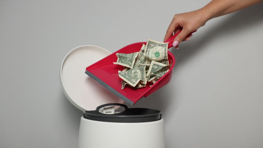 Woman throwing dollar banknotes from dustpan into trash bin near light wall indoors closeup
