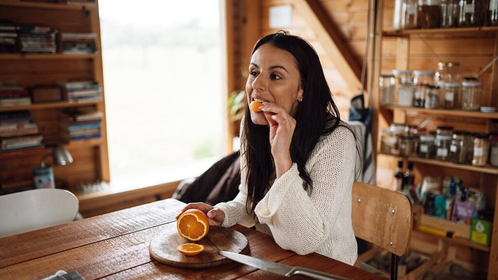A woman sits at a wooden table in a kitchen. She cuts an orange and takes a piece to eat. Sunlight shines through the window, creating a warm setting.