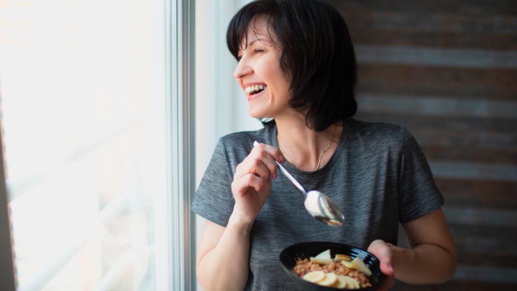 Adult fit slim woman eating breakfast at home. Take care about health and wellness. Senior female person having breakfast meal in bowl. Looking at window.