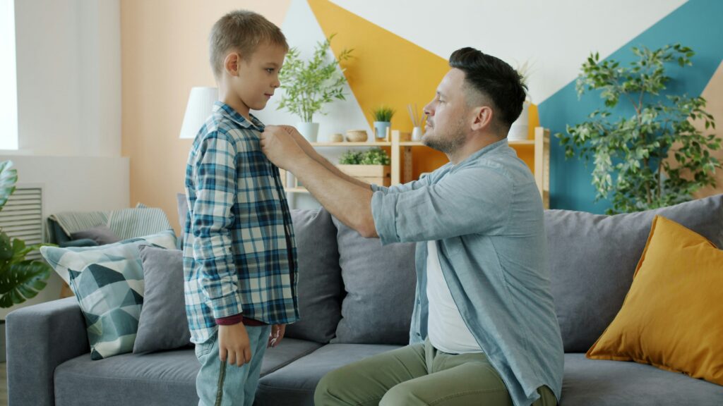 Father buttons son's plaid shirt while sitting on couch.