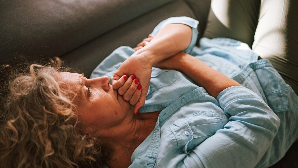 A woman with curly hair lies on a couch, deep in thought with her hand clasped to her mouth. Natural light fills the room, creating a warm and cozy atmosphere.