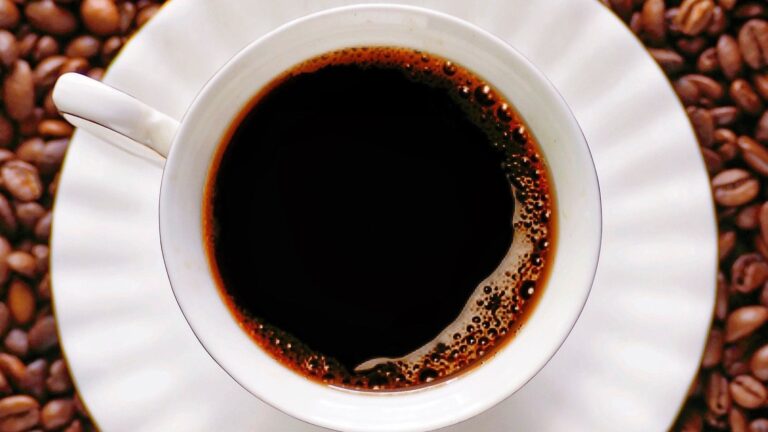 A white cup filled with coffee placed on roasted coffee beans, viewed from above.