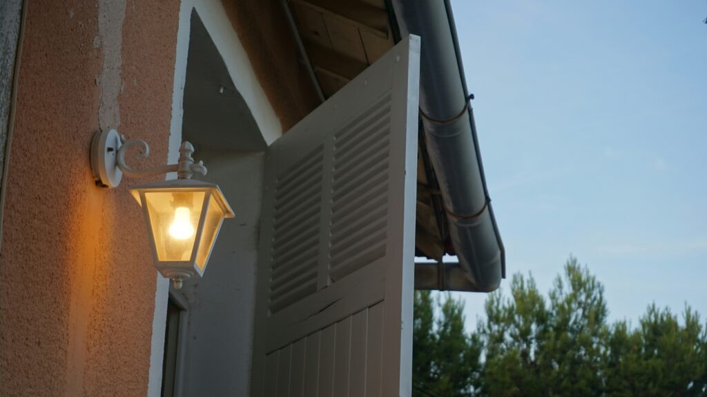 Outdoor lamp illuminates open white shutters on building.