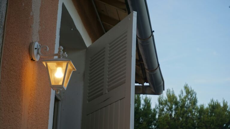 Outdoor lamp illuminates open white shutters on building.