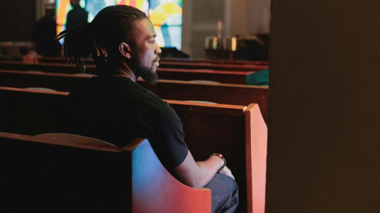 Man sitting in church pews looking towards stained glass.
