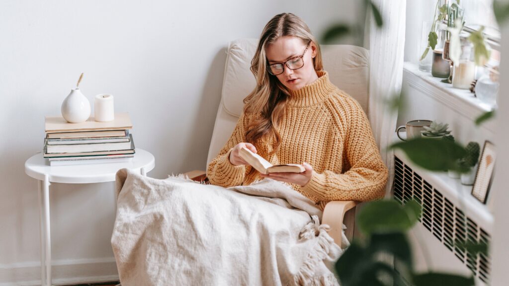 Young female student in trendy sweater and glasses sitting in white armchair with plaid near table with books and candle while reading book