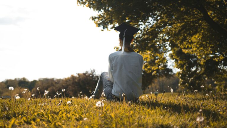 person in white shirt sitting on green grass field with black mortar cap on head