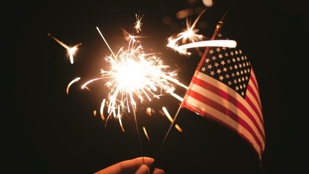 A hand holding a bright sparkler and a small American flag at night