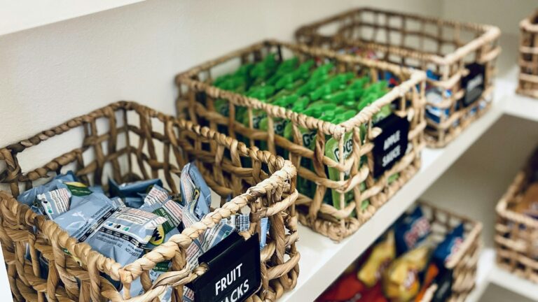 Woven baskets with labels on white pantry shelves filled with various snacks