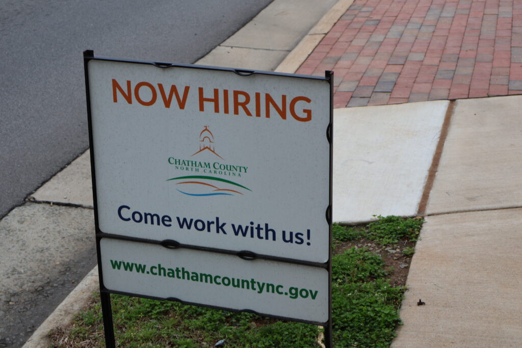 A "Now Hiring, Come work with us" sign on a sidewalk