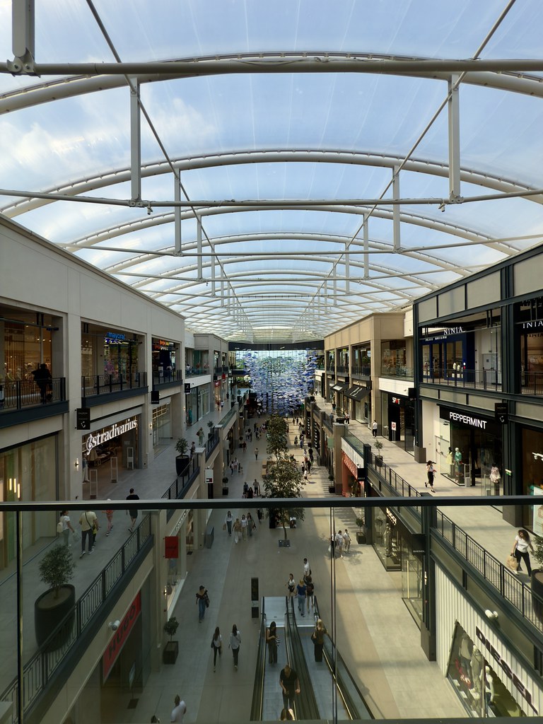 A busy indoor shopping mall with a glass ceiling and storefronts
