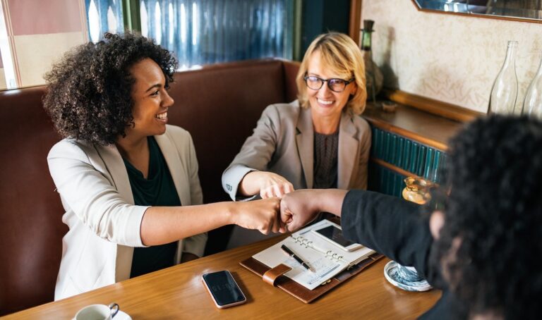 Three businesswomen fist-bumping at a meeting table, illustrating workplace friendship