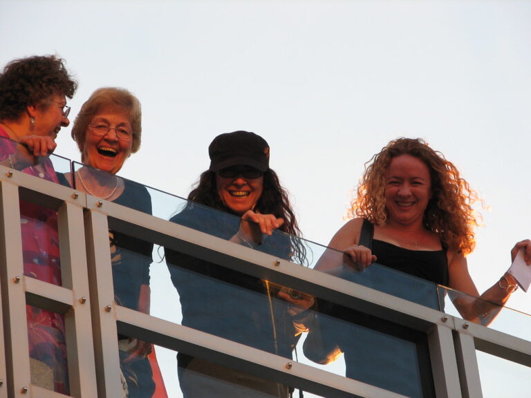 Four women laughing together at a railing in golden-hour light